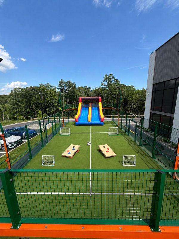 Enclosed turf play space with small soccer goals, cornhole boards and an inflatable slide.